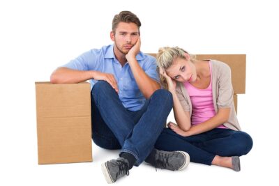 Unhappy young couple sitting beside moving boxes on white backgr